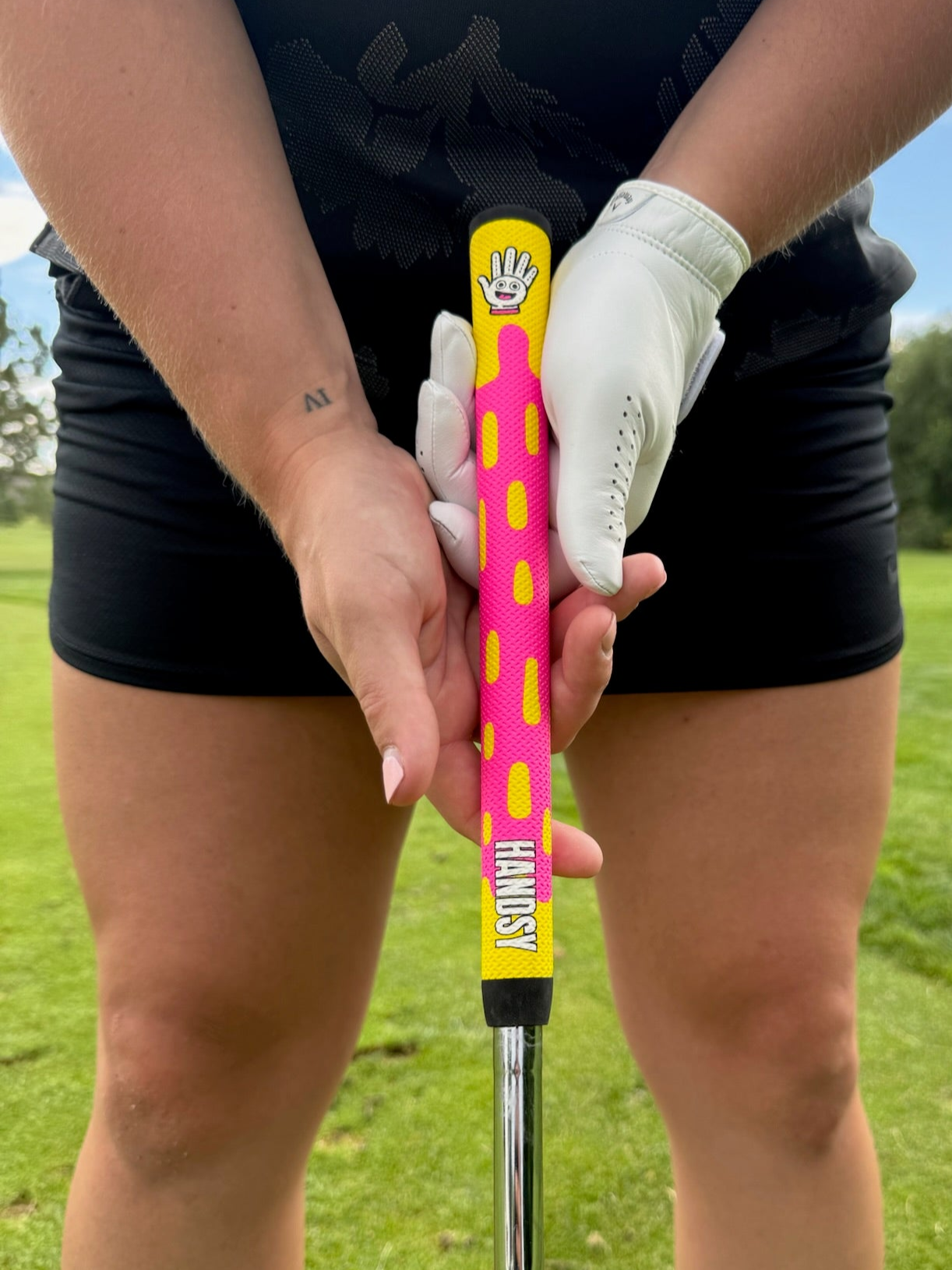 Person holding a golf club with a pink and yellow grip on a golf course.